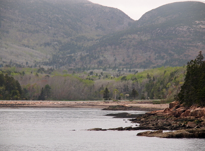 [The mountains rise high around the cove with many green evergreens on the hillside and many bare spots which are probably trees waiting for leaves to appear. There is a flat section of land prior to the rise of the hills.]